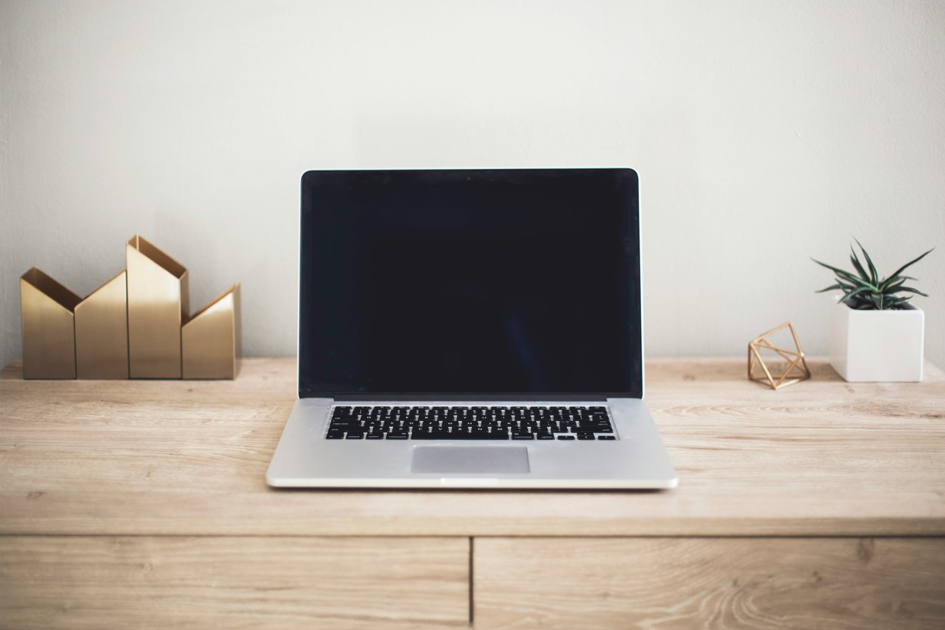 An open laptop on a wooden desk beside a brass document rack and a small potted plant